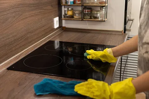 Person cleaning a black electric cooktop with a sponge and yellow gloves.